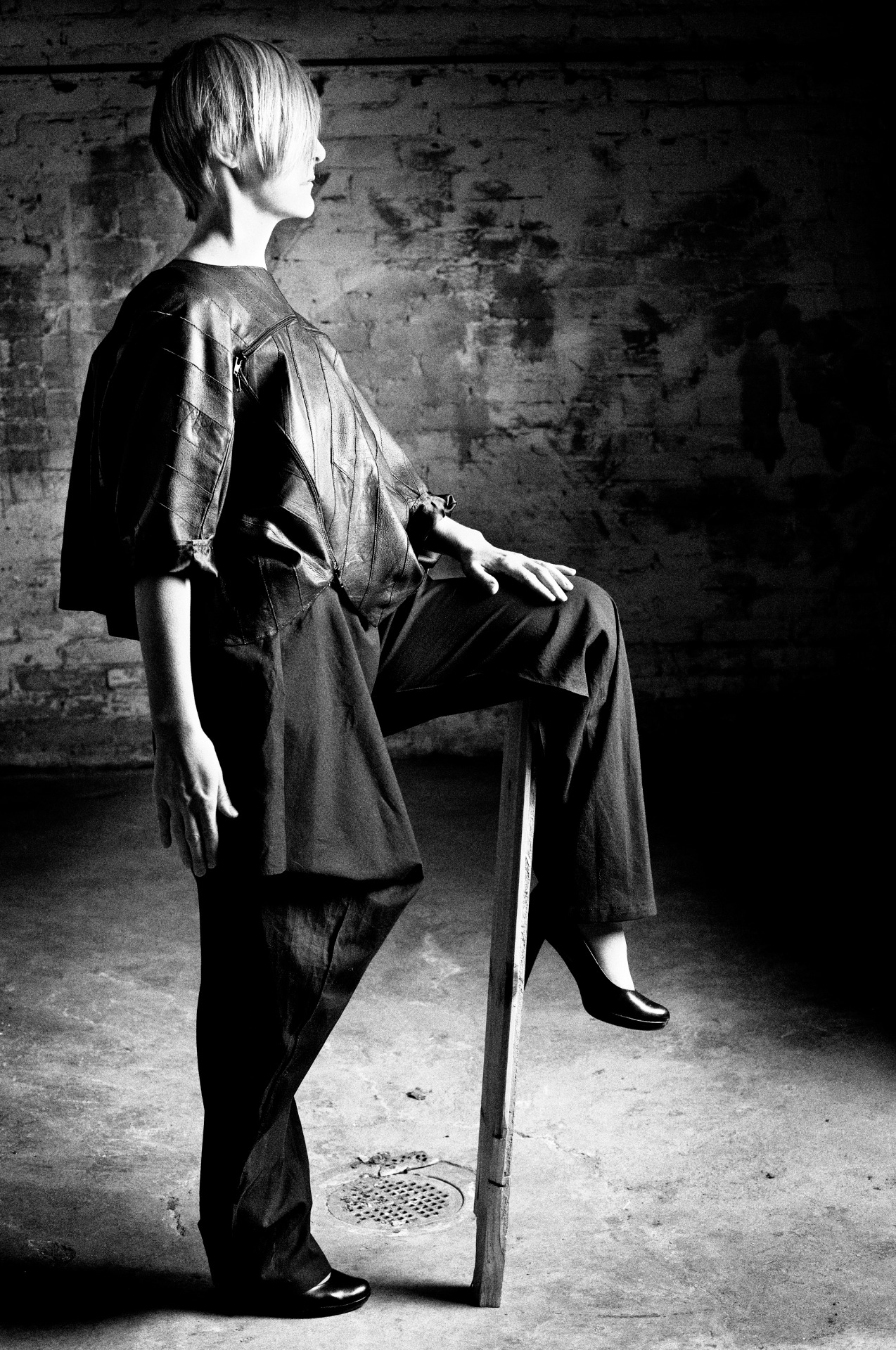 Henkilö seisoo sivuttain jalka korokkeella yllään yhdestä kappaleesta valmistettu nahkatakki ja housut, jotka muodostavat epäsymmetrisen kokonaisuuden.